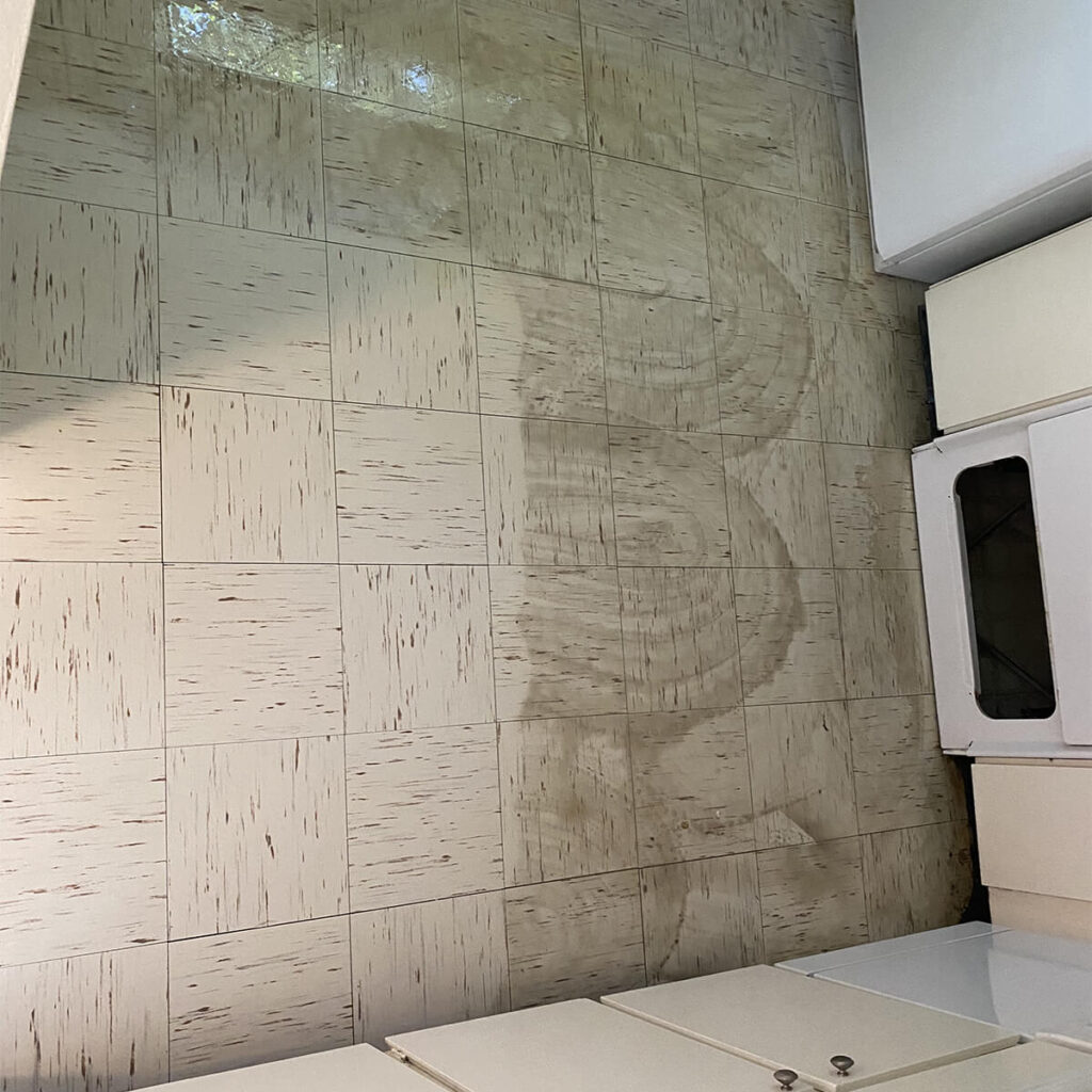 Sea Breeze A tiled floor showing a visible streak of dirt in front of white cabinets and a washing machine, highlighting the need for thorough floor cleaning. Floor And Window Cleaning
