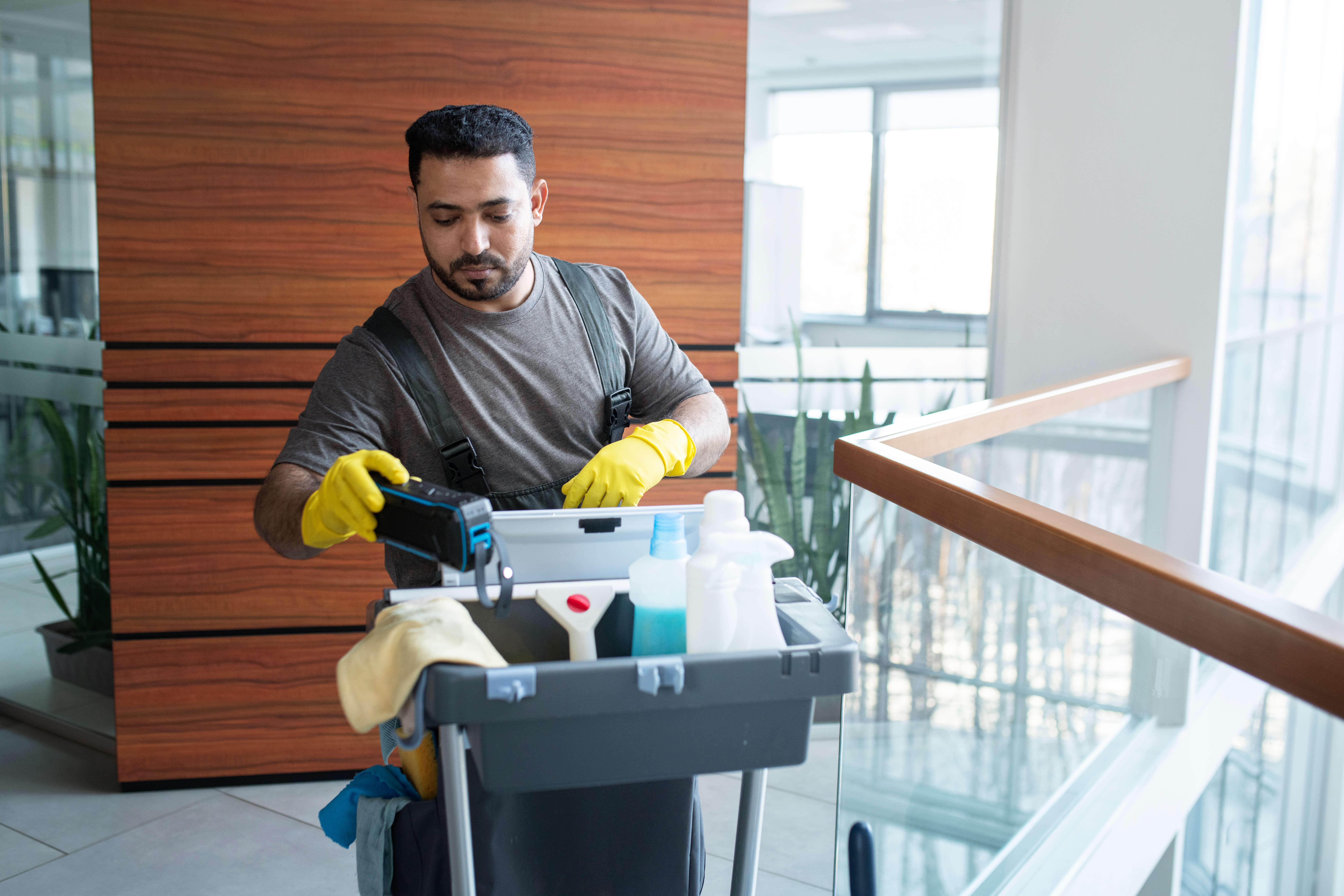 Sea Breeze A janitor wearing gloves and a gray shirt organizes floor cleaning supplies on a cart in a modern office hallway, ensuring everything from mops to Sea Breeze-scented sprays is perfectly arranged. Floor And Window Cleaning