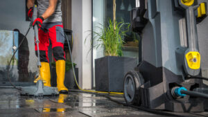 Sea Breeze A person wearing yellow boots and red pants power washes an outdoor patio, standing near a large pressure washer machine. A potted plant is visible in the background near a glass door, as they skillfully tackle both floor cleaning and window cleaning with the refreshing sea breeze in the air. Floor And Window Cleaning