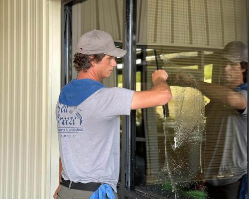 Sea Breeze A man wearing a grey shirt and cap meticulously engages in window cleaning, wielding a squeegee and other tools outside a building, as the sea breeze gently rustles around him. Floor And Window Cleaning